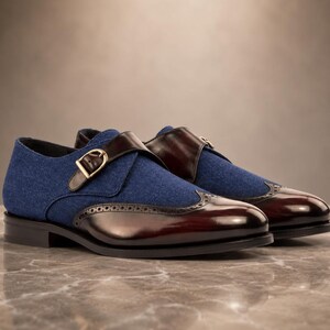 May include: A pair of monk strap shoes featuring a blue fabric upper and a polished dark brown leather toe cap with brogue detailing. The shoes include a single strap across the vamp secured by a gold-toned metal buckle and a low black heel.