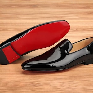 May include: Black patent leather loafers with a glossy finish. One shoe is upright, showcasing a bright red sole. The other shoe is upside down, revealing the red sole and heel. The shoes are on a light brown wooden surface.