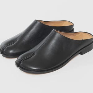 May include: A pair of black leather slip-on shoes with a split-toe design. The shoes have a low heel and a smooth, matte finish. The design is a unique take on a classic shoe style.