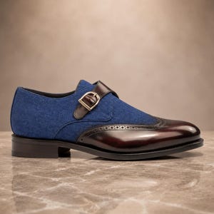 May include: A single monk strap shoe featuring a blue fabric upper and a polished dark burgundy leather toe cap with brogue detailing. The shoe includes a brown leather strap with a gold-toned metal buckle and a low black stacked heel.