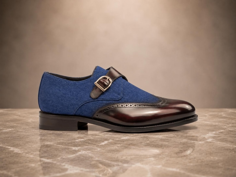 May include: A single monk strap shoe featuring a blue fabric upper and a polished dark burgundy leather toe cap with brogue detailing. The shoe includes a brown leather strap with a gold-toned metal buckle and a low black stacked heel.