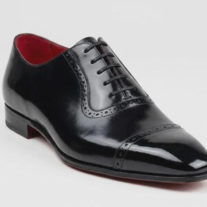 May include: A black leather oxford dress shoe with a glossy finish. The shoe features a red interior, a cap toe, and decorative perforations. The laces are black, and the sole is red.