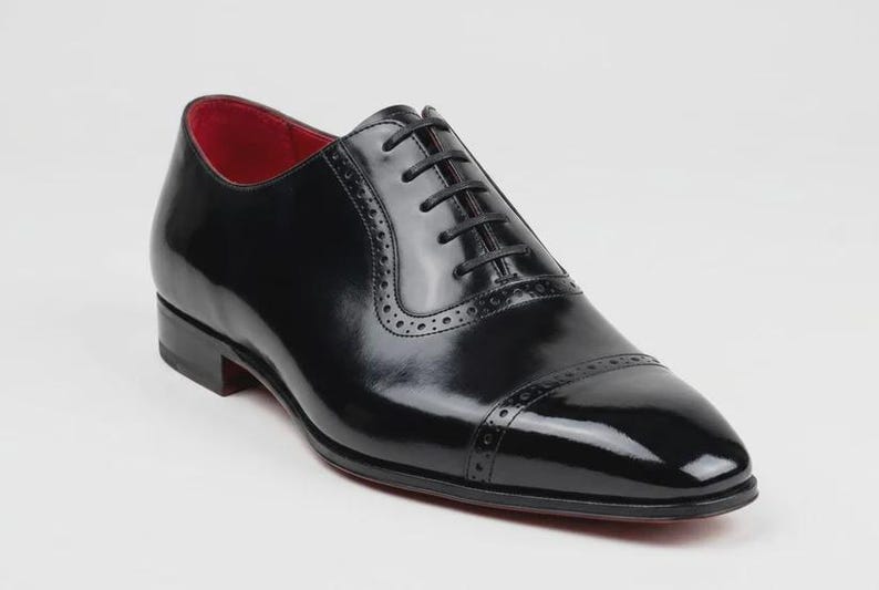 May include: A black leather oxford dress shoe with a glossy finish. The shoe features a red interior, a cap toe, and decorative perforations. The laces are black, and the sole is red.