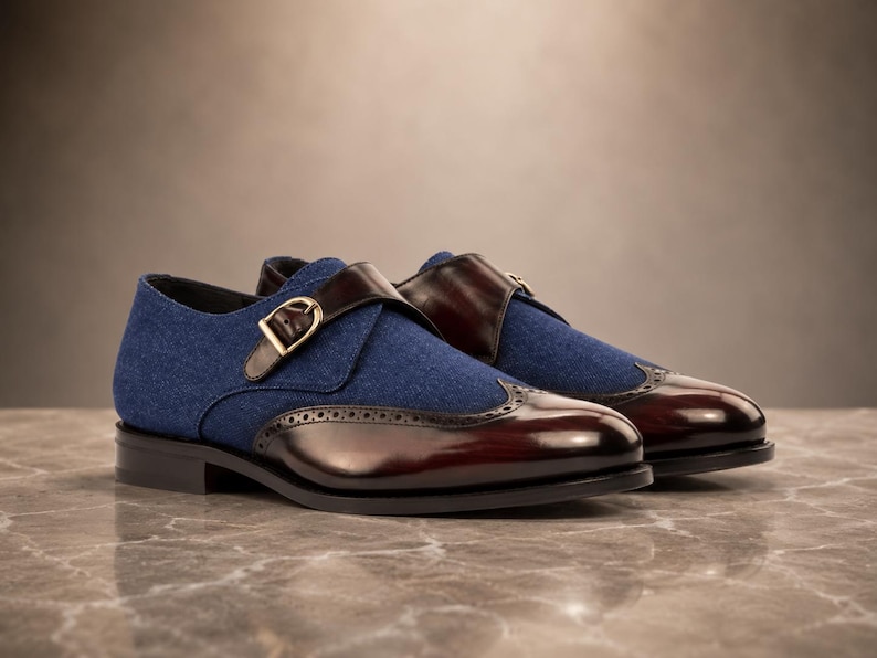 May include: A pair of monk strap shoes featuring a blue fabric upper and a polished dark brown leather toe cap with brogue detailing. The shoes include a single strap across the vamp secured by a gold-toned metal buckle and a low black heel.