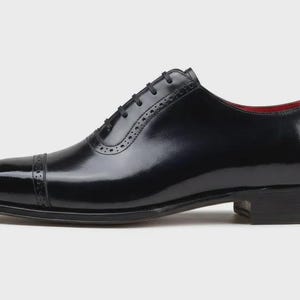 May include: A single black leather dress shoe with a glossy finish. The shoe features a cap toe, decorative perforations, and a lace-up closure. The interior lining is red. The shoe is designed for formal occasions.