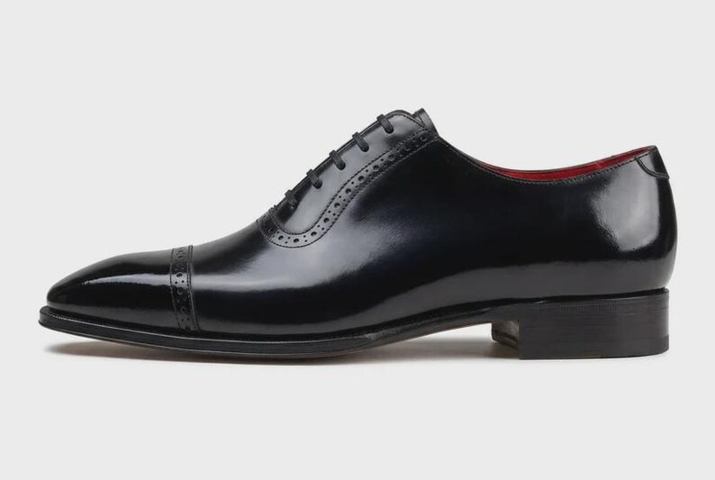 May include: A single black leather dress shoe with a glossy finish. The shoe features a cap toe, decorative perforations, and a lace-up closure. The interior lining is red. The shoe is designed for formal occasions.