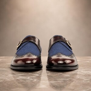 May include: A pair of monk strap shoes featuring a deep burgundy polished leather toe cap with brogue detailing, a blue fabric vamp, and a dark brown leather strap across the top secured with a gold-toned metal buckle on the side of each shoe.