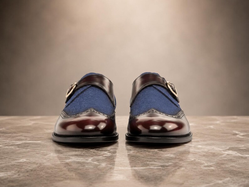 May include: A pair of monk strap shoes featuring a deep burgundy polished leather toe cap with brogue detailing, a blue fabric vamp, and a dark brown leather strap across the top secured with a gold-toned metal buckle on the side of each shoe.