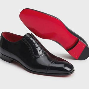 May include: Black leather dress shoes with red soles. The shoes have a classic Oxford design with a cap toe and lace-up closure. The interior is red. One shoe is upright, and the other is angled to show the red sole.