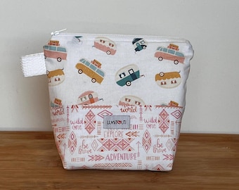 May include: A small, white zippered pouch with a repeating pattern of colorful camper trailers. The bottom half features a geometric design with the words "EXPLORE" and "ADVENTURE". A white fabric loop is attached to the side.