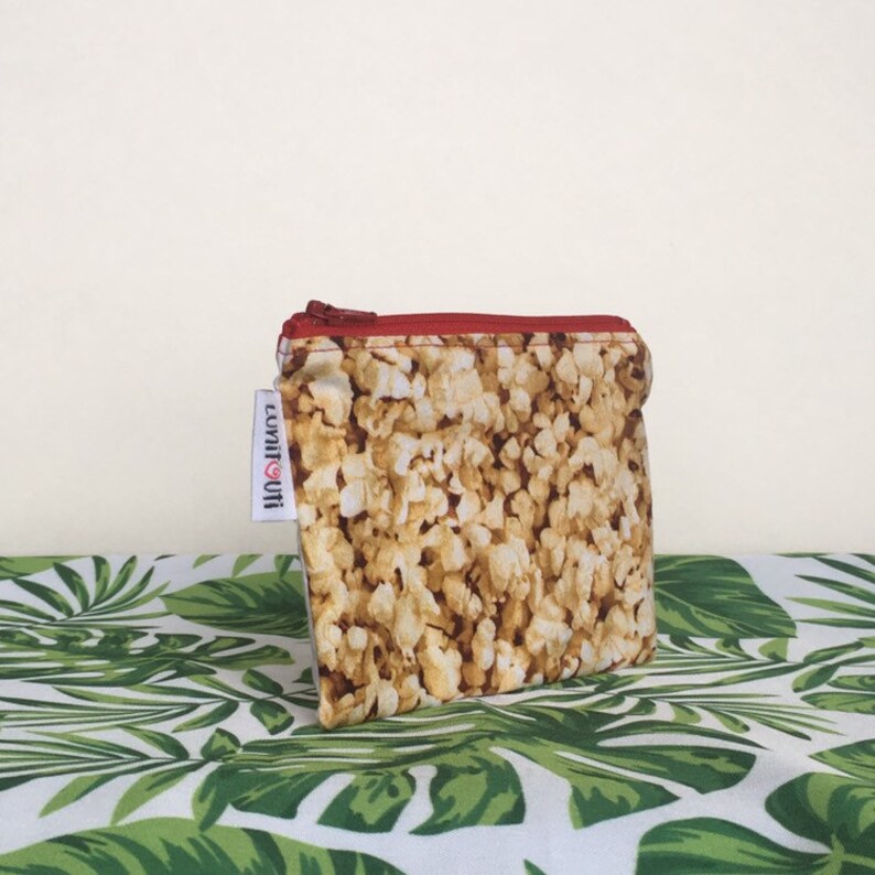 May include: A small zippered pouch with a red zipper and a popcorn print. The pouch is on a green and white tropical leaf patterned fabric. The text "lovin' it!" is on the side of the pouch.