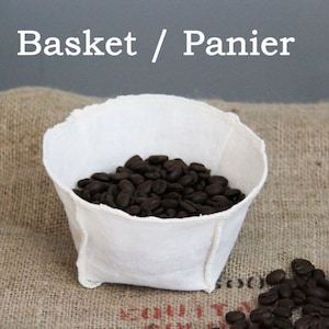 May include: White fabric coffee filter basket filled with dark brown coffee beans. The basket is sitting on a brown hessian surface. Basket / Panier