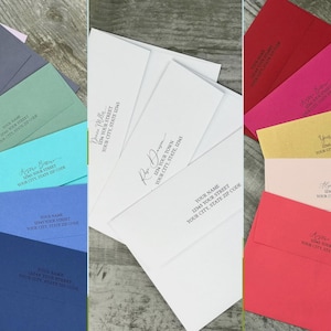 May include: A collection of envelopes in various colors, including blue, teal, gray, and white. Each envelope displays a name and address in a handwritten style. The envelopes are arranged in a fan-like pattern.