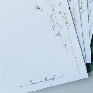 May include: White stationery with delicate watercolor floral illustrations and the name "laura benda" in elegant script. The stationery is paired with green envelopes, creating a cohesive and natural aesthetic.