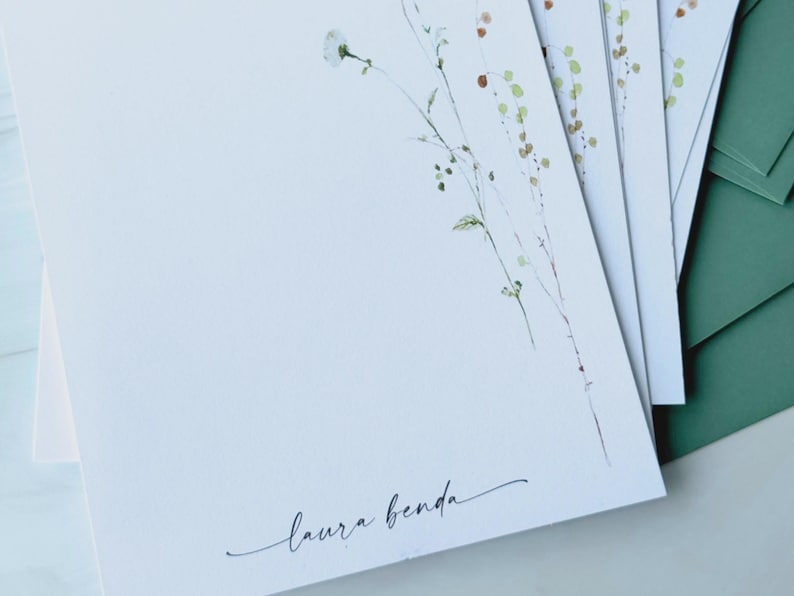 May include: White stationery with delicate watercolor floral illustrations and the name "laura benda" in elegant script. The stationery is paired with green envelopes, creating a cohesive and natural aesthetic.