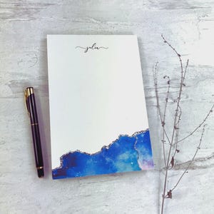 Può includere: Un blocco note bianco con un design ad acquerello blu e viola e bordi glitterati dorati. La parola "Julia" è scritta in corsivo in alto. Una penna nera e dorata è appoggiata accanto al blocco note.