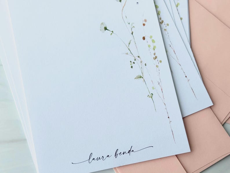 May include: A stack of white stationery with a delicate floral design in green and brown. The name "laura benda" is written in cursive at the bottom. Pink envelopes are visible in the background.