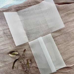 May include: Two unfolded, translucent white paper sheets and one folded paper sheet are arranged on a textured, light brown fabric. A small sprig of dried plant material with leaves is placed near the paper.