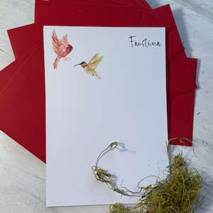 May include: White stationery with watercolor illustrations of a red cardinal and a hummingbird, with the name "Faustina" in script. The stationery is paired with red envelopes and a sprig of dried greenery.