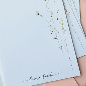 May include: A stack of white stationery with a delicate floral design in the upper right corner. The stationery has the name "laura benda" in cursive script. Pink envelopes are visible in the background.