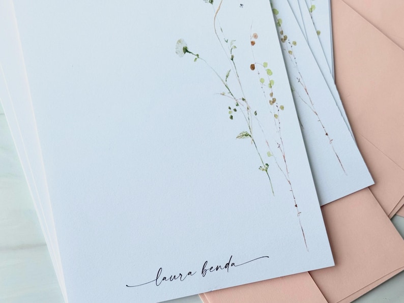 May include: A stack of white stationery with a delicate floral design in the upper right corner. The stationery has the name "laura benda" in cursive script. Pink envelopes are visible in the background.