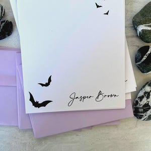 May include: White stationery featuring black bat silhouettes and the name "Jasper Brown" in script. Lilac envelopes and decorative stones are also visible.