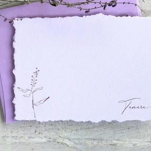 May include: A handmade lavender stationery set featuring a deckled-edge card with a delicate botanical illustration and the name "Tamara" in cursive. The card rests on a matching lavender envelope, with dried sprigs in the background.
