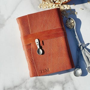 May include: A brown leather journal with a strap closure and the initials "TSM" embossed on the cover. A set of measuring spoons and a bowl of oats are also in the image, suggesting a theme of cooking or journaling.