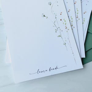 May include: A stack of white stationery with delicate floral illustrations and the signature "laura benda" in script. Green envelopes are visible in the background, suggesting a stationery set. The floral design features light green and brown accents.