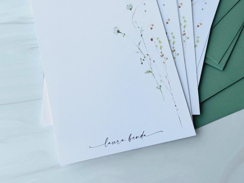 May include: A stack of white stationery with delicate floral illustrations and the signature "laura benda" in script. Green envelopes are visible in the background, suggesting a stationery set. The floral design features light green and brown accents.