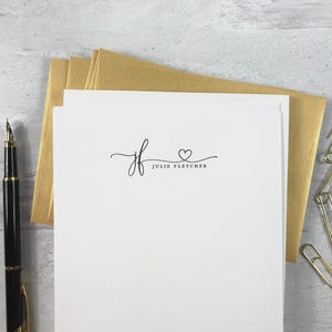 Puede incluir: Papelería blanca con un bolígrafo negro, sobres dorados y clips. La papelería presenta las iniciales "jf" con un corazón y el nombre "Julie Fletcher" en una escritura delicada. El bolígrafo es negro con detalles dorados.