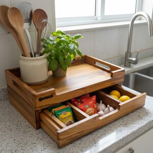 May include: A wooden kitchen organizer with a tray top and a pull-out drawer. The drawer contains lemons, garlic, and spice packets. A small potted plant and utensils in a ceramic jar sit on top. The organizer is made of light brown wood.