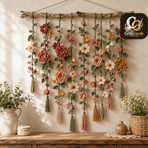 May include: A wall hanging featuring multiple vertical strands of crocheted flowers and leaves in shades of pink, orange, and cream, suspended from a natural wooden branch with a jute cord. Each strand ends in a small, matching yarn tassel.