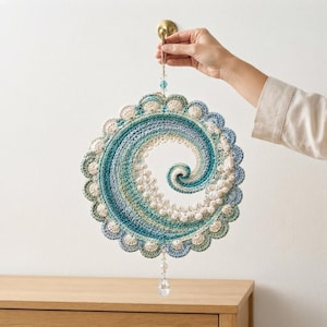 May include: A circular crochet wall hanging featuring a spiral wave pattern in shades of teal, blue, and cream. The piece is edged with a scalloped border and accented with small white beads along the spiral. A clear crystal drop hangs from the bottom.
