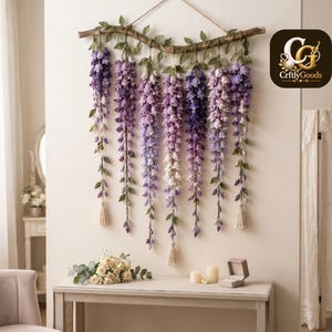 May include: A wall hanging featuring crocheted wisteria flowers in shades of purple, lavender, and white, suspended from a natural wooden branch with green crocheted leaves. The piece includes small cream-coloured tassels and a logo reading CrftlyGoods.