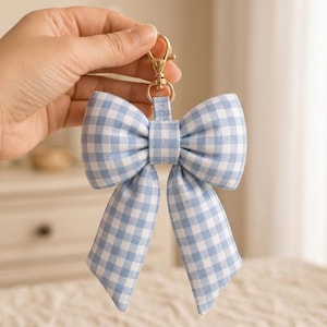 May include: A fabric keychain featuring a large bow with a light blue and white gingham check pattern. The bow is attached to a gold-toned metal swivel lobster clasp, providing a secure way to attach the accessory to bags or keys.
