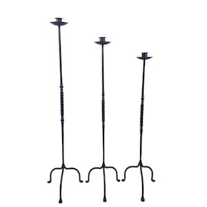 Op de afbeelding: Een set van drie zwarte metalen vloerkandelaars van verschillende hoogtes. Elke kandelaar heeft een slanke verticale steel met een gedraaid middengedeelte, een ronde kaarshouder aan de bovenkant en een driepootvoet met gebogen poten.