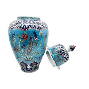 Puede incluir: Un tarro de cerámica con tapa, que presenta un fondo azul claro decorado con intrincados patrones florales en rojo, azul oscuro, blanco y verde. El diseño incluye tulipanes y claveles estilizados, con bordes azul oscuro en la parte superior y la base.