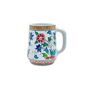 May include: A white ceramic mug featuring a hand-painted floral pattern with red, blue, and green petals and leaves. The mug has a thick handle on the right side and decorative brown and orange bands with intricate line details at the top and base.