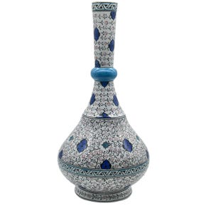 Op de afbeelding: Een hoge, bolvormige vaas met een smalle hals en een uitlopende voet, voorzien van een witte achtergrond versierd met ingewikkelde zwarte bloemmotieven, kleine rode accenten en grotere blauwe bloembladmotieven, afgewerkt met een geometrische rand.