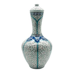 Op de afbeelding: Een hoge keramische vaas met bijpassend deksel, voorzien van een witte basis versierd met ingewikkelde zwarte bloemranken, kleine rode accenten en een opvallende blauwe band rond de schouder met witte geometrische patronen en een meander-rand.