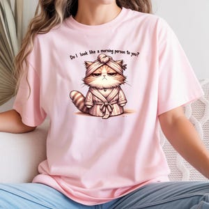 Puede incluir: Camiseta rosa claro con un gato de dibujos animados gruñón con una bata y un turbante. El texto en la camiseta dice: "¿Te parezco una persona mañanera?"