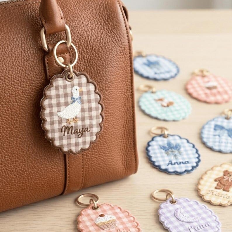 May include: A brown leather handbag with a gold-colored metal ring holding a personalized keychain. The keychain features a gingham pattern, a white duck illustration, and the name "Maya". Other keychains with different designs are visible.