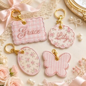 May include: Four decorative keychains in pink and gold. One is a rectangle with the name "Grace", another is a flower shape with "Lily", an oval with a floral pattern, and a butterfly shape. All have gold key rings.