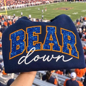 May include: A navy blue sweatshirt featuring the text BEAR in large, blue glitter block letters with orange outlines, and the word down in white cursive script underneath. The garment is held up against a blurred background of a football stadium.