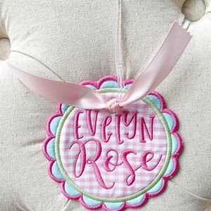 May include: A personalized, embroidered ornament with the name "Evelyn Rose" in pink script. The ornament features a pink and white gingham pattern, a scalloped edge, and a light pink ribbon for hanging.