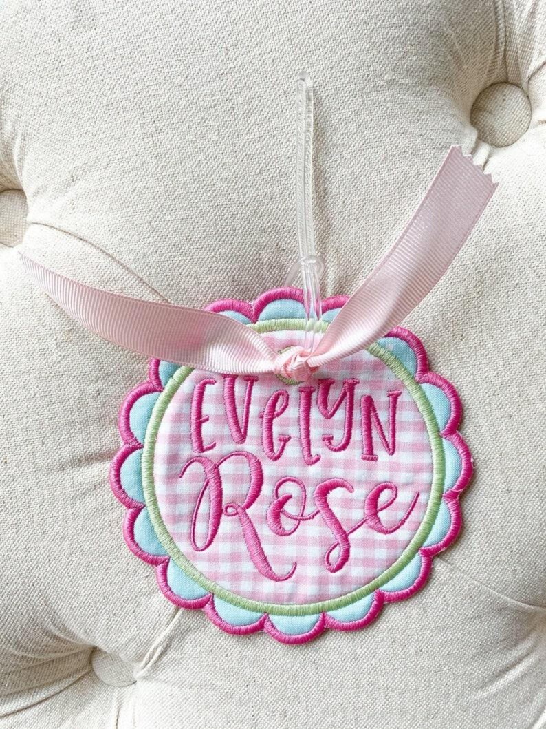 May include: A personalized, embroidered ornament with the name "Evelyn Rose" in pink script. The ornament features a pink and white gingham pattern, a scalloped edge, and a light pink ribbon for hanging.