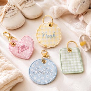 May include: A collection of personalized baby keychains in various shapes and colors. The keychains include a pink heart with "Ella", a yellow scalloped edge with "Noah", a blue circle with "Sophie", and a green rectangle. Gold-tone hardware is attached.