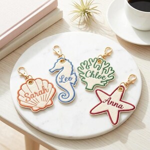 May include: Four embroidered keychains with gold-colored clasps. The keychains are shaped like a seashell, seahorse, coral, and starfish. Each keychain has a name embroidered on it: Sarah, Leo, Chloe, and Anna. The keychains are on a white marble surface.