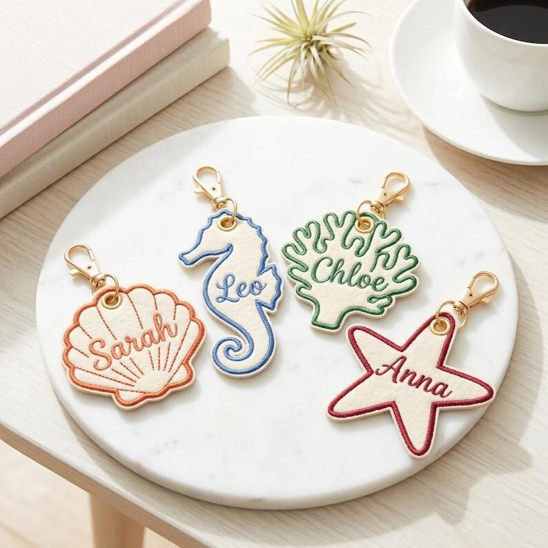 May include: Four embroidered keychains with gold-colored clasps. The keychains are shaped like a seashell, seahorse, coral, and starfish. Each keychain has a name embroidered on it: Sarah, Leo, Chloe, and Anna. The keychains are on a white marble surface.